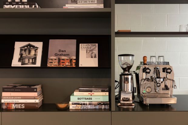 A refined built-in shelving and coffee station with dark matte cabinetry, curated art and design books, and a stainless-steel espresso machine and grinder on the right. The backdrop features a white painted masonry or block wall, while the shelving creates a warm, contemporary library-bar atmosphere.