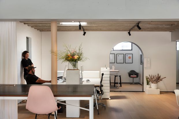 A bright contemporary office interior with an open-plan workspace, dark communal desk, pale pink office chair, white desks and chairs, and a small meeting or work area visible through an arched opening. The space features exposed timber ceiling battens, pale walls, warm timber flooring, black track lighting, and indoor plants, creating a refined, minimalist workplace atmosphere.
