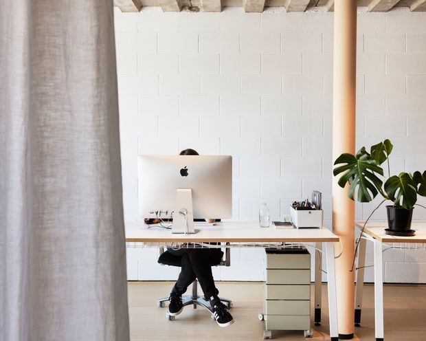Bright minimalist home office with a white-painted concrete block wall, exposed timber ceiling beams, pale timber flooring, and a simple white desk. An iMac, rolling office chair, mobile drawer unit, shelving, and a large potted monstera are visible. A warm-toned structural column and sheer curtain frame the workspace.