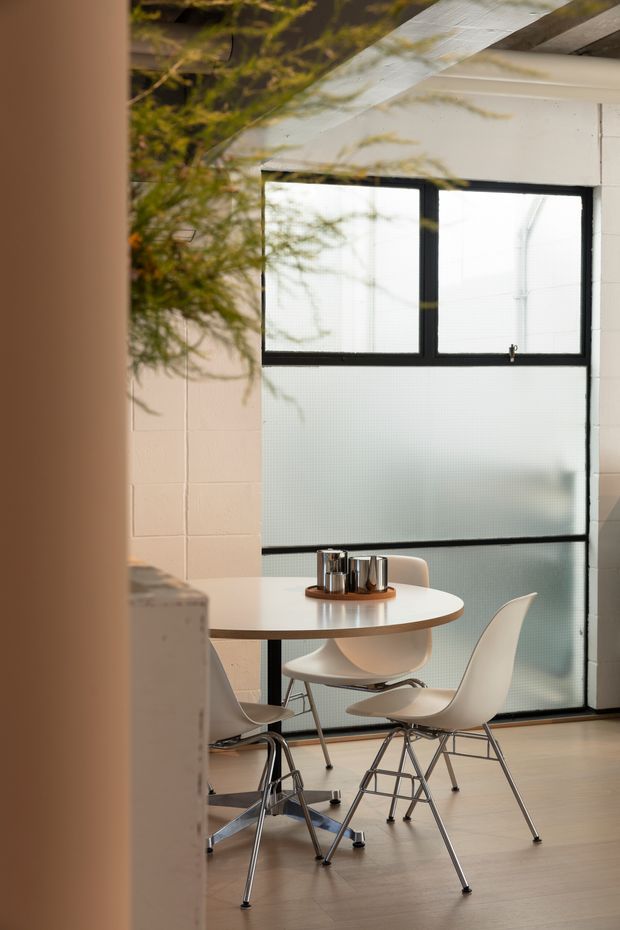 A softly lit contemporary dining area viewed through a partial doorway or wall opening. The space features a round white dining table with a small metallic centrepiece, sculptural white chairs with chrome-finished legs, pale timber flooring, white tiled walls, and large black-framed frosted glazing. A sloped translucent roof or clerestory-like glazed structure brings in diffused daylight, while greenery is visible near the opening.