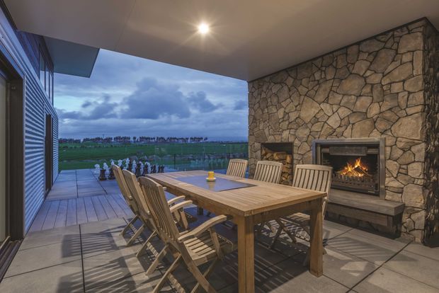 Covered outdoor dining and entertaining area with a substantial natural-stone feature wall, built-in fireplace, timber dining table and folding timber chairs. The open side overlooks a rural landscape, while large-format paved flooring and a recessed ceiling with downlighting create a sheltered outdoor room.