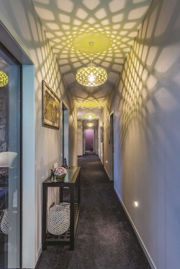 A long, narrow residential hallway with dark carpet, pale textured walls, framed artwork, and a slim console table with decorative objects. Multiple ornate perforated pendant lights create dramatic geometric patterned shadows across the ceiling and walls. Doorways and glimpses of adjacent rooms line the corridor, with subtle low-level wall lighting adding atmosphere.