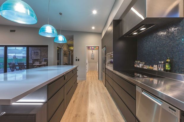 Contemporary galley kitchen with a large pale island, dark flat-panel cabinetry, stainless-steel appliances and benchtops, and a dark blue mosaic-tile splashback. Pendant lights hang above the island, while full-height glazing connects the kitchen to an outdoor area.