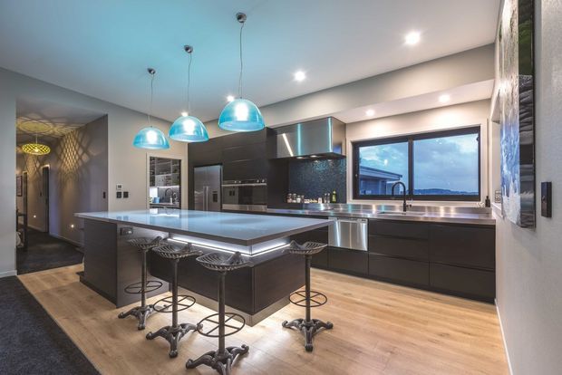 Contemporary open-plan kitchen with a large pale stone island, dark flat-panel cabinetry, integrated stainless-steel appliances, and four industrial-style bar stools. Three turquoise glass pendant lights hang above the island, while a wide window above the sink provides an exterior view. The space has timber flooring, recessed ceiling lighting, and a dark tiled or textured feature wall near the entry.