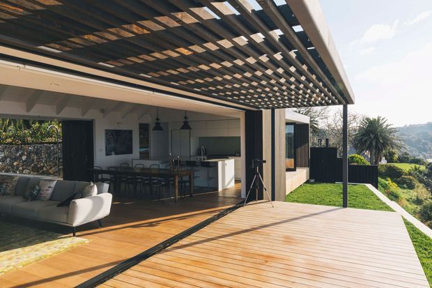 An open-plan indoor-outdoor living and dining area opening onto a large timber deck. The space features a contemporary kitchen with island, dining table, sofa seating, expansive sliding glass doors, and a dramatic overhead timber pergola creating a patterned shadow across the deck. Dark vertical screening and timber cladding contrast with light walls, while a landscaped sloping site is visible beyond.