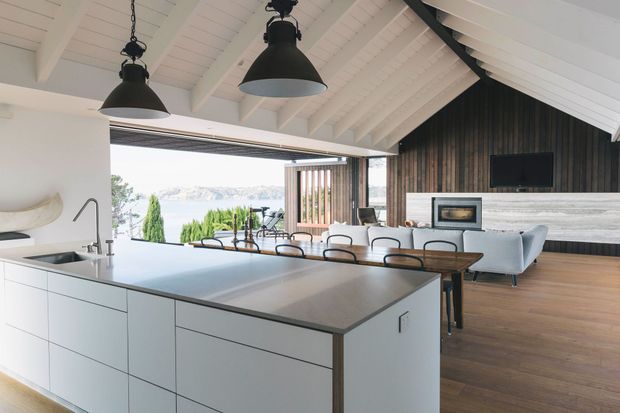 Bright open-plan kitchen and living area with a large white island, integrated sink and tap, dark benchtop, dining table, contemporary seating, and a fireplace set into a dramatic dark timber feature wall. The room has a white vaulted timber-lined ceiling with exposed beams, oversized black pendant lights, extensive open glazing, and views toward a landscaped waterfront or hillside setting.