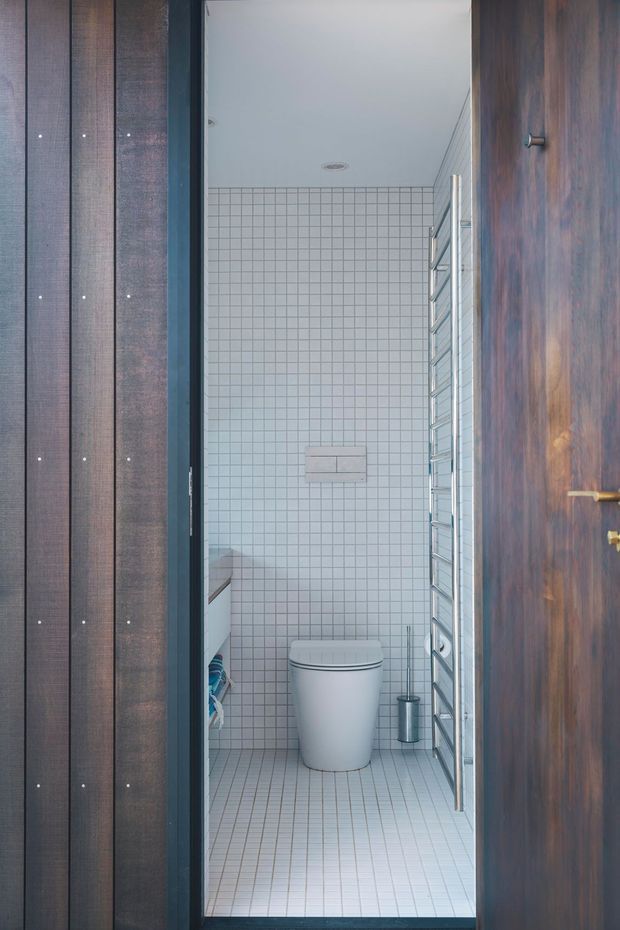 Compact contemporary bathroom viewed through an open timber door, featuring white small-format grid tiles on the walls and floor, a wall-hung toilet, brushed metal heated towel rail, concealed flush plate, and minimal white fixtures. Dark timber wall cladding and a dark timber door frame the space.