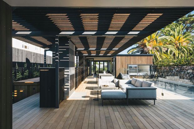 A spacious covered outdoor living and entertaining area with a timber deck, large modular outdoor sofa, dining or serving furniture, built-in barbecue cabinetry, and a slatted pergola overhead. Glazed balustrades open the space toward a pool or landscaped area, while a timber-clad structure and tropical planting are visible beyond. The design combines dark structural framing with warm timber surfaces and strong sunlight-and-shadow patterns.