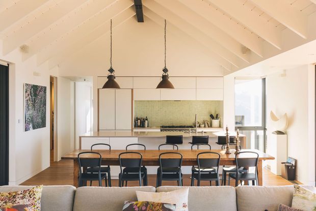 Bright open-plan kitchen and dining area viewed from the living room. The space features a warm timber floor, white built-in cabinetry, a pale green small-tile splashback, a timber kitchen island with integrated seating, a long timber dining table with black chairs, and a vaulted white-painted timber ceiling with exposed beams. Large windows and a glazed door provide abundant natural light. Black pendant lights and dark window/door frames add contrast.