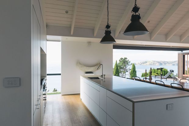 A bright contemporary kitchen with a large white island, integrated cabinetry, timber flooring, and a white painted exposed-beam ceiling. Black pendant lights hang above the island, while wide open glazing connects the kitchen to an outdoor dining area and a scenic waterfront landscape.