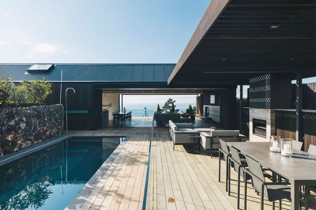 A contemporary coastal outdoor entertaining area with a rectangular swimming pool, extensive timber decking, covered dining and lounge zones, and dark-clad pavilion structures. The open-plan space frames a distant ocean view, while a stone retaining wall, outdoor furniture, and integrated sheltered areas complete the setting.