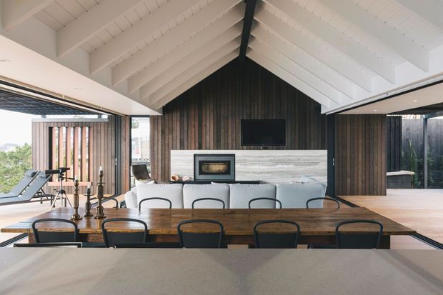 Open-plan dining and living area with a long timber dining table in the foreground, a pale upholstered sofa, and a central fireplace and television wall. The room has dramatic white pitched timber rafters with a dark central structural beam, dark vertical timber wall cladding, extensive glazing, and a glimpse of exercise equipment at the side.
