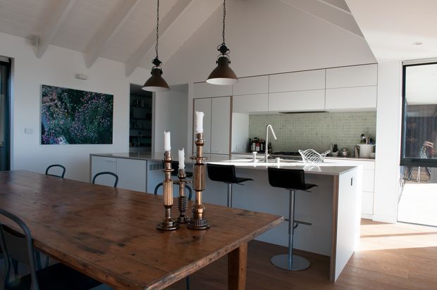 Bright open-plan kitchen and dining area with a large timber dining table in the foreground, a white island with integrated sink and bar seating, pale cabinetry, mirrored splashback, and exposed white-painted ceiling beams. Pendant lights hang above the island, while glazed doors bring in natural light.