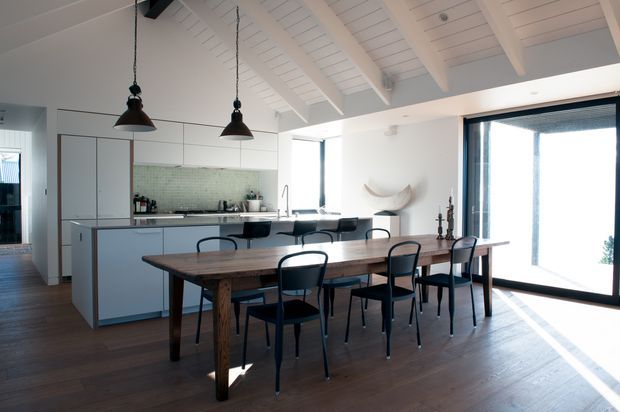A bright open-plan kitchen and dining area with a long dark timber dining table and black dining chairs in the foreground. The kitchen has pale cabinetry, a pale green tiled splashback, dark benchtops, and a central island with stools. Large glazed openings bring in strong natural light, while a white pitched ceiling with exposed beams and white lining boards creates an airy, coastal feel. Black pendant lights hang above the dining area, and dark timber flooring runs throughout.