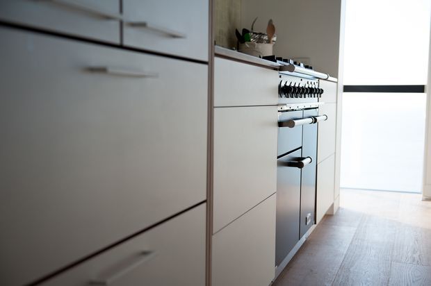 Close-up view of a sleek contemporary kitchen with extensive white handleless cabinetry, integrated drawers, a built-in oven, and a pale timber floor. A bright window or glazed door is visible at the end of the narrow kitchen run.
