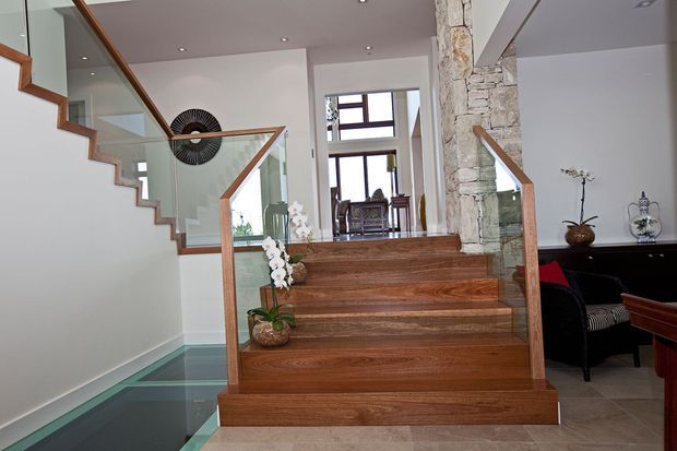 Interior view of a contemporary split-level stairway featuring broad timber stairs, clear glass balustrades with timber handrails, white walls, and a prominent rough-stone structural column. A lower-level glazed floor area and a furnished living or dining space are visible beyond the stairs.