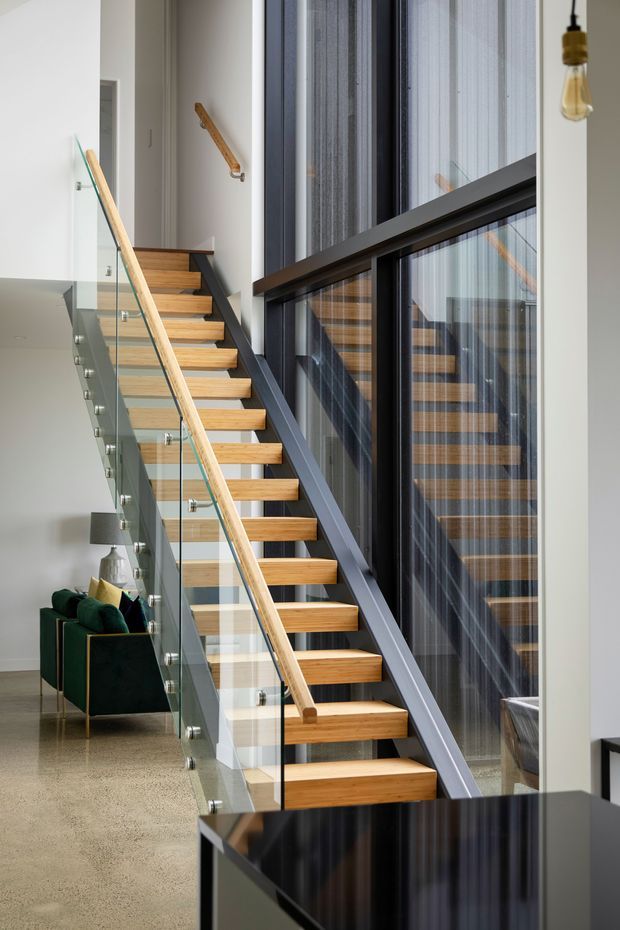 Bright contemporary interior focused on an open timber staircase with a dark painted steel stringer, timber handrail, and clear glass balustrade. Full-height glazing runs alongside the stair, reflecting the steps and bringing in abundant natural light. A green upholstered armchair and side table sit in the lower living area, with a glossy dark benchtop or cabinet in the foreground.