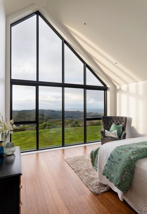 A bright bedroom with a dramatic floor-to-ceiling triangular and rectangular black-framed window wall overlooking a green rural landscape and distant hills. The room has pale angled walls and ceiling, warm timber flooring, a tufted dark armchair, a bed with a botanical green quilt, a soft area rug, and a dark bedside cabinet with decorative objects. Strong natural light creates geometric shadows across the room.