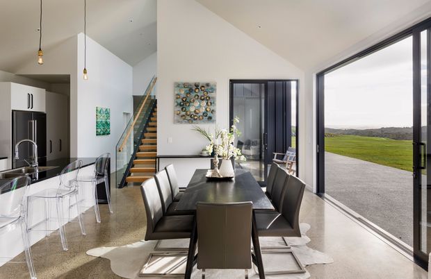 Contemporary open-plan dining and kitchen interior with a long dark dining table and upholstered chairs, a white kitchen with island and transparent dining chairs, polished concrete flooring, and a timber-and-metal staircase. Large black-framed sliding glass doors open to a paved terrace and expansive green landscape, creating a bright, airy connection to the outdoors.