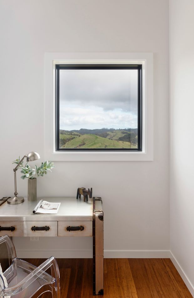 A compact, light-filled home office corner with a white-painted desk or cabinet, vintage-style dark metal hardware, a chrome desk lamp, a small vase of greenery, and transparent acrylic seating. A large black-framed window provides a view of rolling green hills. The room has pale walls, white trim, and warm dark timber flooring.