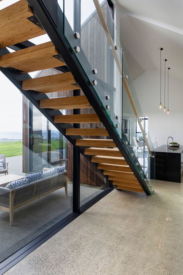 A bright contemporary interior featuring a sculptural open-riser staircase with warm timber treads, dark steel stringers, and a glass balustrade. Full-height glazing opens to a rural landscape, while a compact black kitchen island and pendant lights are visible in the background. The polished speckled concrete floor and exposed timber elements create a refined modern aesthetic.