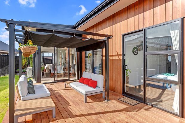 A bright contemporary outdoor living area with a timber deck beside a vertically clad timber exterior wall. The deck includes white outdoor sofas, red cushions, a black metal pergola with retractable shade canopy, glass doors, and a hanging planter. A bedroom or interior space is visible through the glazing.