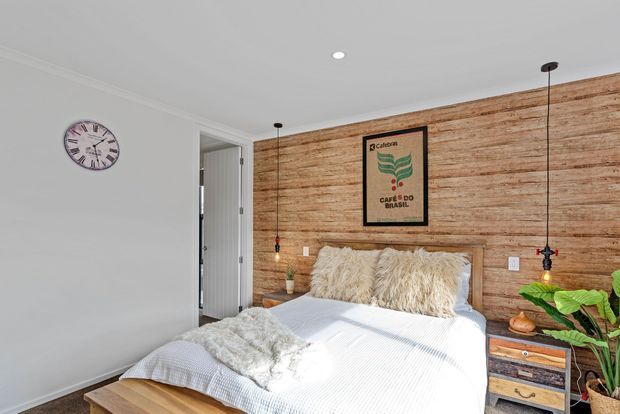 A bright bedroom with a double bed against a full-height reclaimed timber feature wall. The room has white walls and bedding, shaggy cream cushions, a light timber bedside table, hanging pendant lights, a wall clock, framed artwork, and a leafy indoor plant.