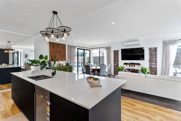Open-plan contemporary kitchen and living area with a large white island, dark cabinetry, integrated appliances, timber flooring, and pendant lighting. The space includes a dining area, sectional sofa, wall-mounted television, indoor plants, and expansive glazed doors/windows leading to an outdoor area.