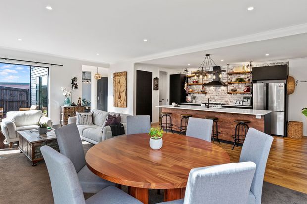 Open-plan interior combining a dining area, living room and kitchen. A large round timber dining table with upholstered chairs sits in the foreground, while the kitchen has a timber island, open shelving, stainless-steel appliances, dark cabinetry and pendant lighting. The living area includes pale sofas and armchairs beside large glazed doors opening to an outdoor deck. The space is bright, warm and contemporary with some traditional detailing.