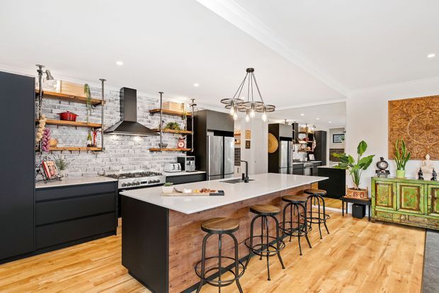 Open-plan industrial-style kitchen and living area with a large island, white stone benchtop, black cabinetry, exposed white brick splashback, timber flooring, open timber shelving, stainless-steel appliances, and black metal bar stools. A decorative green console and wall artwork are visible in the adjoining living space.