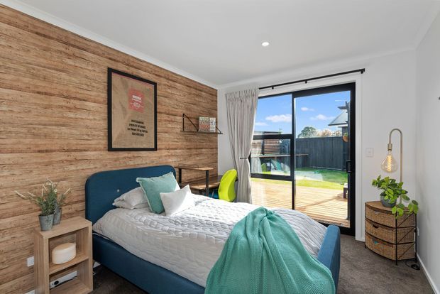 A bright, contemporary bedroom with a teal upholstered bed, layered white and teal bedding, and a full-height timber feature wall. Black-framed sliding glass doors open to a timber deck and fenced outdoor area, while a small bedside table, framed artwork, open shelving, woven storage, plants, and a pendant lamp add warmth and personality.