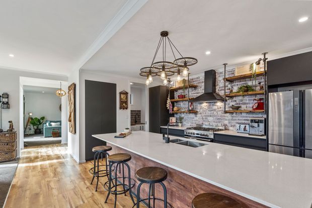 Bright open-plan kitchen with a large white waterfall-edge island and timber-clad base, industrial-style stools, dark cabinetry, open metal shelving, stainless-steel appliances, and a white-and-grey brick backsplash. A decorative multi-arm pendant hangs above the island, while timber flooring and an adjacent hallway with a glimpse of a living area are visible.