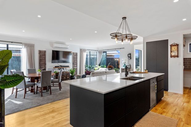 Open-plan contemporary kitchen, dining and living area with a large dark island, white benchtop, timber flooring, dining table, television feature wall, sofas and expansive windows. A linear pendant chandelier hangs above the island, while white walls and recessed lighting create a bright, spacious interior.