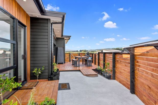 A contemporary residential outdoor terrace and entry courtyard with dark horizontal cladding, warm timber wall panels, timber decking, concrete paving, black-framed glazing, raised planter pots, and a compact outdoor dining setting. Timber privacy fencing encloses the space, with a bright open sky and distant landscape beyond.