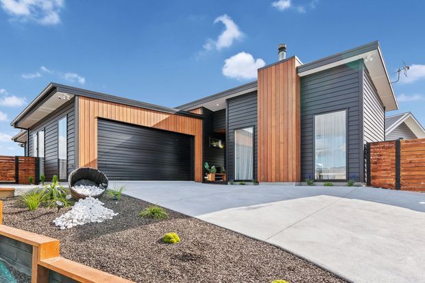 Contemporary single-storey house exterior with a flat-roofed, asymmetrical form. The facade combines dark horizontal cladding with warm vertical timber cladding, large glazed openings, and an integrated double garage. A broad concrete driveway and low-maintenance gravel garden with sculptural landscaping occupy the foreground, while timber fencing encloses the site.