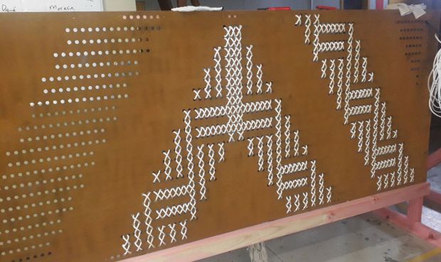 A close-up of a large brown wooden board mounted on a simple pink-painted frame. The board is covered with rows of small white pegs or fasteners arranged in geometric, stepped and zigzag patterns, resembling a hands-on construction, weaving, or educational display. Part of a workshop or classroom setting is visible in the background.