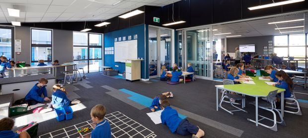 Bright, open-plan school learning commons or classroom with children working at tables and on the carpet. The space features large windows, glass-partitioned rooms, blue accent walls, dark carpet flooring with colourful geometric inlays, suspended rectangular LED lights, white acoustic ceiling tiles, and flexible contemporary school furniture.