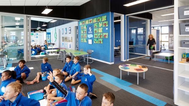 Bright contemporary school learning commons or classroom area with children seated on a carpeted floor in blue uniforms. The space has extensive internal glazing, blue accent walls, colourful educational artwork, collaborative tables, stools, open shelving, and suspended rectangular fluorescent lighting. Multiple adjoining learning spaces are visible through glass partitions.