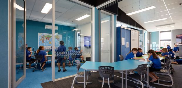 Bright contemporary commercial learning space with glazed meeting or classroom rooms, turquoise-blue walls and carpet, rows of student seating, collaborative tables, and people working or studying. The space has a suspended acoustic ceiling with recessed fluorescent lighting, extensive internal glazing, and a blue-and-white educational fit-out.