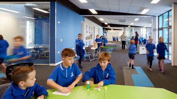 A bright, contemporary school or educational common area with children in blue uniforms working and socialising around colourful tables. The open-plan interior has glazed meeting or classroom partitions, blue accent walls, a suspended acoustic ceiling with recessed lights, carpeted flooring, and activity areas extending into the background.