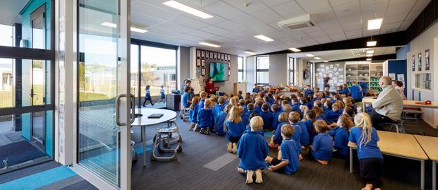 A bright, contemporary school multipurpose or assembly room viewed through open glazed doors. A large group of children in blue uniforms sit on dark carpet facing a presentation area, with long light-wood tables and benches, a kitchenette or service counter, extensive glazing, and a suspended acoustic ceiling with recessed lighting. The space has a practical institutional fit-out and strong daylight.
