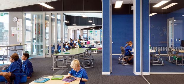 A bright contemporary school or educational interior with an open collaborative dining or study area, blue glass-partitioned rooms, rows of tables and chairs, and students using the space. Dark carpet, blue feature walls, extensive interior glazing, and linear fluorescent-style ceiling lights define the fit-out.