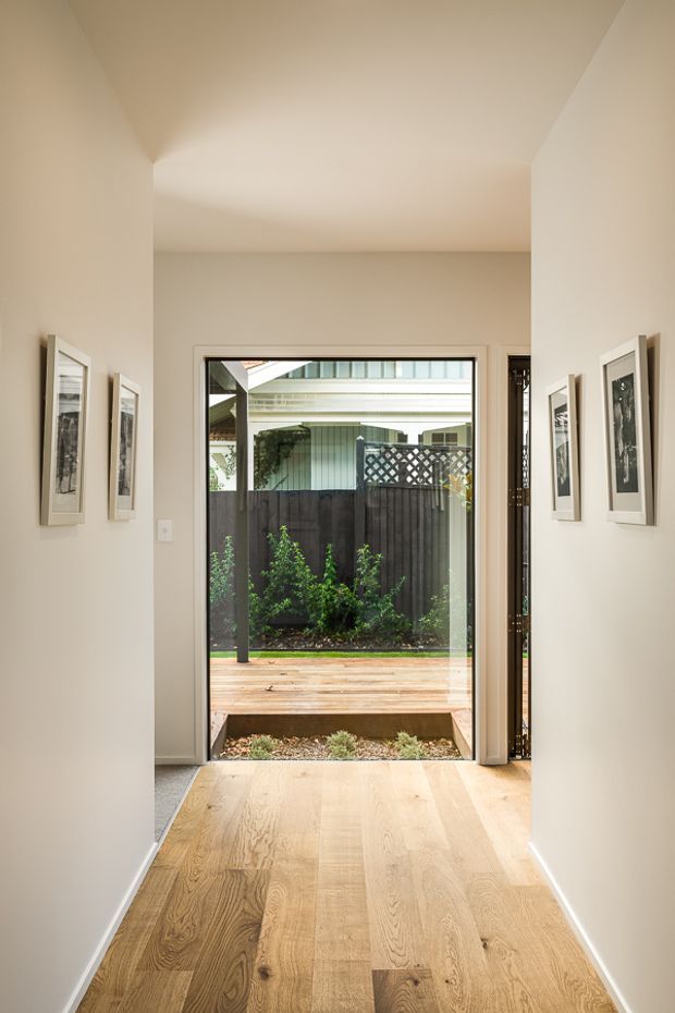 A bright, narrow residential hallway or entry passage with light oak timber flooring, warm off-white walls, and framed artwork arranged along both sides. The passage opens through full-height glazed doors to a small timber deck and landscaped courtyard, with a dark privacy fence and neighbouring structure visible beyond. The space has a clean, contemporary architectural character and strong indoor-outdoor connection.