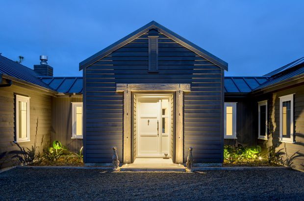 A symmetrical evening view of a low, single-storey residential building with a dark blue-grey horizontal weatherboard facade, pitched gable roof and central illuminated entry porch. Warm light glows from the white front door and surrounding windows, with gravel paving and planted beds in the foreground.
