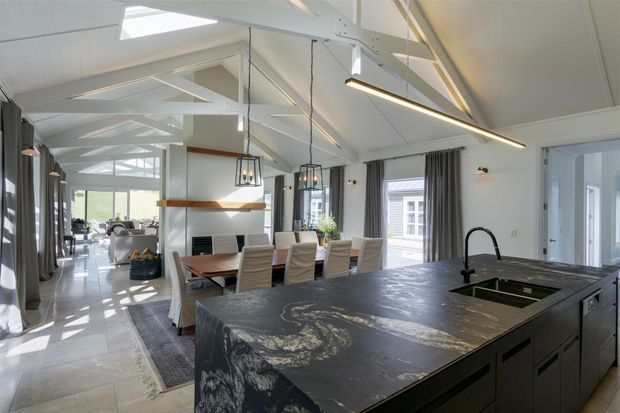 Bright open-plan kitchen and dining area with a large dark stone island and integrated sink in the foreground. The space features white cabinetry and walls, light tiled flooring, exposed painted structural trusses, skylights, pendant lights, and expansive glazing with grey curtains. A dining table and living area extend into the background.