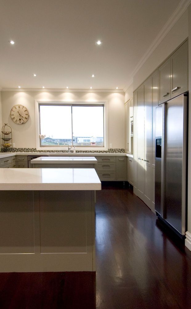A bright, spacious traditional kitchen with a large central island, pale built-in cabinetry, white stone benchtops, stainless-steel refrigerator, dark polished timber flooring, recessed ceiling lights and decorative cornices. A patterned mosaic tile splashback runs beneath the window and along the rear benchtop.