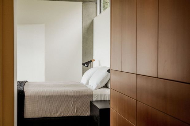 A compact, minimalist bedroom viewed through a doorway, featuring a neatly made bed with neutral textured bedding, a black bedside table and wall-mounted reading light. Full-height warm timber built-in cabinetry dominates the right side, while a concrete-toned wall and white surfaces create a restrained contemporary interior.