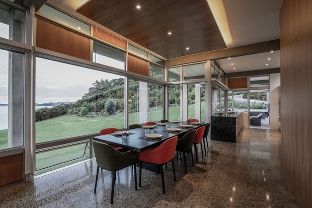 A refined open-plan dining area with a long dark dining table and red-and-black chairs, surrounded by extensive floor-to-ceiling glazing overlooking a landscaped lawn and distant hills. Warm timber wall and ceiling panels, slim dark window frames, recessed lighting, and polished terrazzo-style flooring create a contemporary, light-filled interior. A kitchen or service area is visible beyond the dining space.