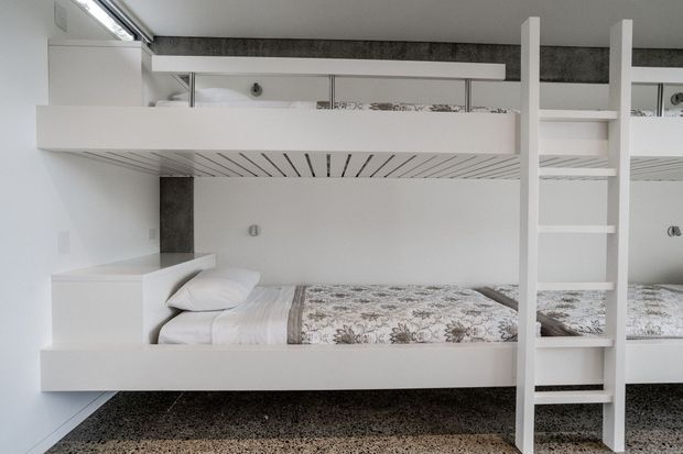 Compact modern bunk bedroom with two built-in sleeping levels, white joinery, integrated ladder, slim horizontal guardrails, and a patterned grey-and-white duvet. The room has a clean monochromatic palette, terrazzo-like flooring, and minimal built-in detailing.