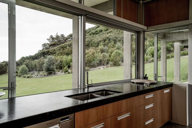 A contemporary kitchen with a long dark stone island and integrated double sink, warm timber cabinetry with streamlined horizontal handles, and expansive floor-to-ceiling glazing. The glazing opens views toward a landscaped sloping garden, while concrete structural columns and reflected cabinetry reinforce the modern architectural character.